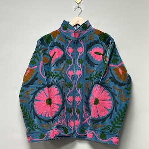 May include: A blue embroidered jacket with a high collar and a zipper closure. The jacket features large pink floral designs, green leaves, and brown accents. The jacket is hanging on a wooden hanger.