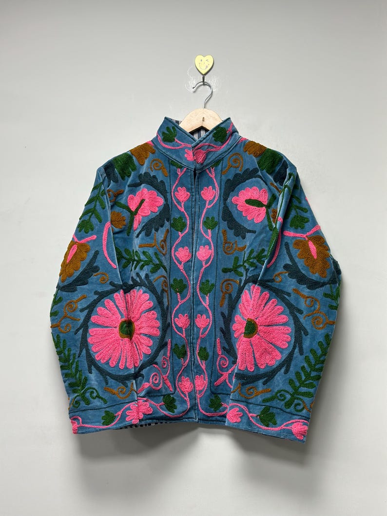 May include: A blue embroidered jacket with a high collar and a zipper closure. The jacket features large pink floral designs, green leaves, and brown accents. The jacket is hanging on a wooden hanger.