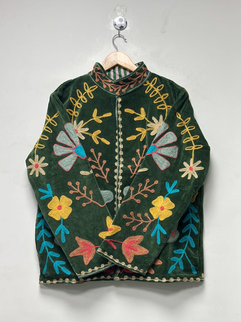 May include: A dark green velvet jacket featuring a high collar and long sleeves. The jacket is decorated with vibrant, multi-colored floral and leaf embroidery in shades of yellow, light blue, pink, and brown, with a striped lining visible at the neck.