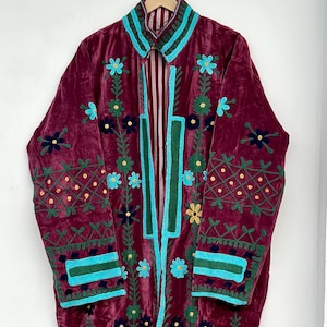 May include: A long-sleeved burgundy velvet coat featuring intricate embroidery in shades of turquoise, dark green, and yellow. The garment displays floral motifs, geometric patterns on the sleeves, and a striped lining visible at the collar.