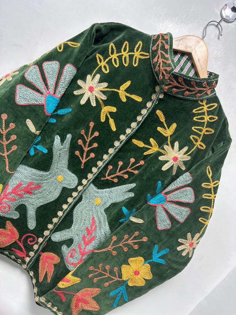 May include: A dark green velvet jacket featuring intricate multi-colored embroidery of rabbits, flowers, and leaves. The garment has a high collar, long sleeves, and a front opening with a decorative stitched border and a striped inner lining.