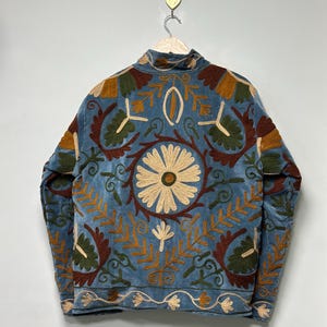 May include: A light blue jacket featuring intricate embroidery in shades of cream, dark brown, and olive green. The back displays a large central daisy motif surrounded by symmetrical floral and leaf patterns, with a decorative border along the hem.