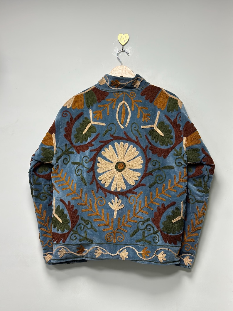 May include: A light blue jacket featuring intricate embroidery in shades of cream, dark brown, and olive green. The back displays a large central daisy motif surrounded by symmetrical floral and leaf patterns, with a decorative border along the hem.