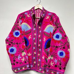 K&ouml;nnte beinhalten: Eine leuchtend pinke Jacke mit aufwendigen Blumenstickereien in Blau-, Rot- und Gr&uuml;nt&ouml;nen. Das Kleidungsst&uuml;ck hat einen Stehkragen, lange &Auml;rmel und ein am Halsausschnitt sichtbares, vertikal gestreiftes Innenfutter mit Knopfleiste.