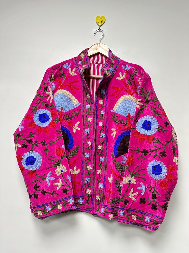 K&ouml;nnte beinhalten: Eine leuchtend pinke Jacke mit aufwendigen Blumenstickereien in Blau-, Rot- und Gr&uuml;nt&ouml;nen. Das Kleidungsst&uuml;ck hat einen Stehkragen, lange &Auml;rmel und ein am Halsausschnitt sichtbares, vertikal gestreiftes Innenfutter mit Knopfleiste.