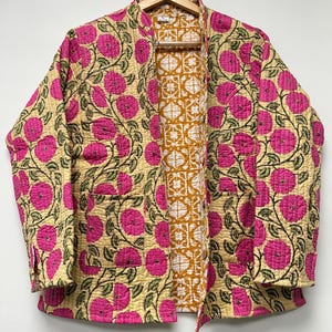 May include: A quilted jacket with a floral design. The jacket has a pale yellow base, adorned with large pink flowers and green foliage. The lining is mustard yellow with a white geometric pattern. It has two pockets and is displayed on a wooden hanger. The jacket is approximately 76 cm long.