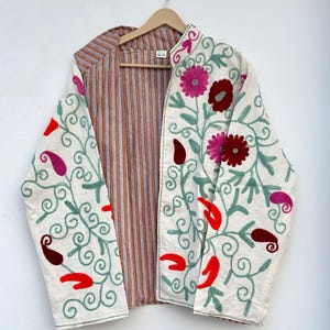 May include: A long-sleeved jacket featuring an off-white base with intricate floral and paisley embroidery in shades of pink, red, and sage green. The interior lining displays a brown and white vertical striped pattern, visible at the open front.