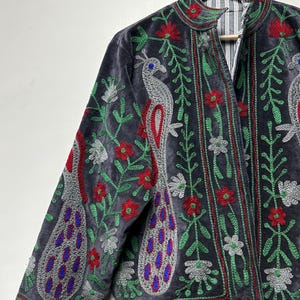 May include: A charcoal gray velvet jacket featuring intricate embroidery of silver peacocks with blue accents, surrounded by red flowers and green vines. The jacket has a high collar and a vertical striped lining visible at the open front neckline.