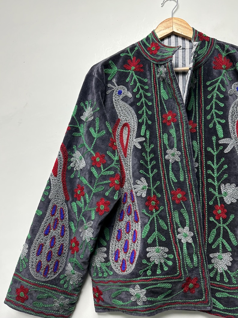 May include: A charcoal gray velvet jacket featuring intricate embroidery of silver peacocks with blue accents, surrounded by red flowers and green vines. The jacket has a high collar and a vertical striped lining visible at the open front neckline.