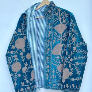 May include: A teal velvet jacket featuring intricate beige floral embroidery on the front and sleeves. The jacket is open, revealing a white and blue vertical striped lining on the interior. It has a high collar and long sleeves.