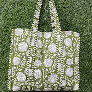 Puede incluir: Un bolso tote verde con un estampado floral blanco. El bolso tiene asas largas y está hecho de tela acolchada. El diseño floral incluye flores grandes y hojas. El bolso está sobre una superficie verde.