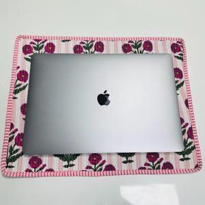 May include: A silver laptop rests on a floral patterned mat. The mat features a pink and white striped background with repeating magenta flower and green leaf designs. A pink border frames the mat.
