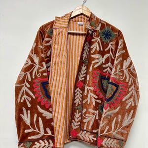 Puede incluir: Una chaqueta de terciopelo marr&oacute;n con intrincados bordados florales en tonos rojo, verde y azul. La chaqueta tiene mangas largas, cuello alto y una parte delantera abierta que revela un forro interior con rayas verticales naranjas y blancas.