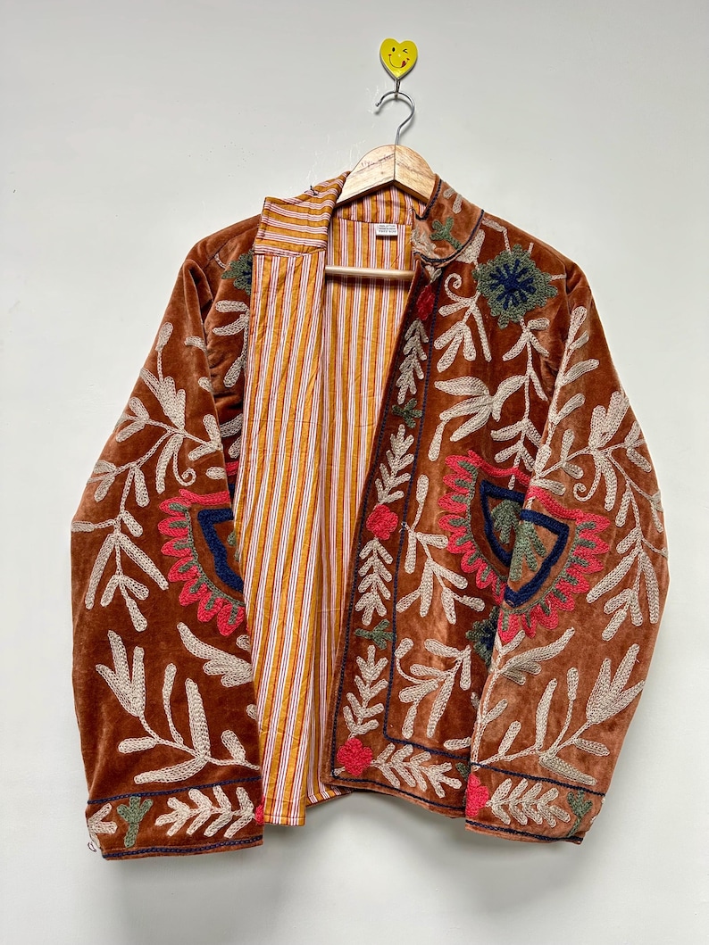 Puede incluir: Una chaqueta de terciopelo marr&oacute;n con intrincados bordados florales en tonos rojo, verde y azul. La chaqueta tiene mangas largas, cuello alto y una parte delantera abierta que revela un forro interior con rayas verticales naranjas y blancas.