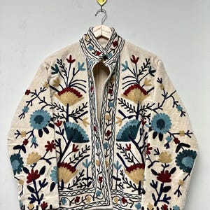 May include: Cream-colored embroidered jacket with a floral design. The jacket features blue, red, and yellow flowers with green stems and leaves. The embroidery is detailed and covers the front and sleeves of the jacket. The jacket is hanging on a wooden hanger.