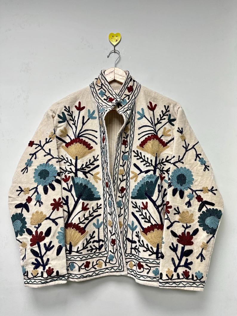 May include: Cream-colored embroidered jacket with a floral design. The jacket features blue, red, and yellow flowers with green stems and leaves. The embroidery is detailed and covers the front and sleeves of the jacket. The jacket is hanging on a wooden hanger.