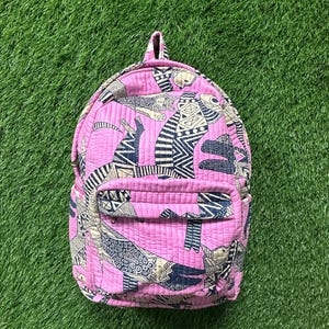 May include: A pink quilted backpack featuring a repeating pattern of black and off-white stylized figures. The backpack has a front pocket and a top handle. It is set against a background of green grass.