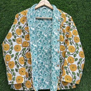 Puede incluir: Chaqueta acolchada reversible con estampado floral. Un lado presenta flores amarillas sobre fondo blanco, mientras que el otro lado tiene un diseño floral turquesa. La chaqueta tiene cierre de botones y está colgada de una percha de madera.