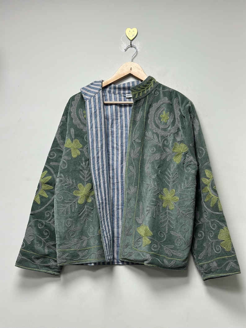 May include: A sage green jacket with floral embroidery and a striped lining. The jacket is hanging on a wooden hanger against a white wall. The embroidery is in a lighter green color.