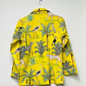 Puede incluir: Camisa amarilla con botones con estampado de tem&aacute;tica selv&aacute;tica. La camisa presenta cebras, leopardos, tucanes y flamencos entre palmeras y otro follaje. La camisa est&aacute; colgada en una pared blanca.