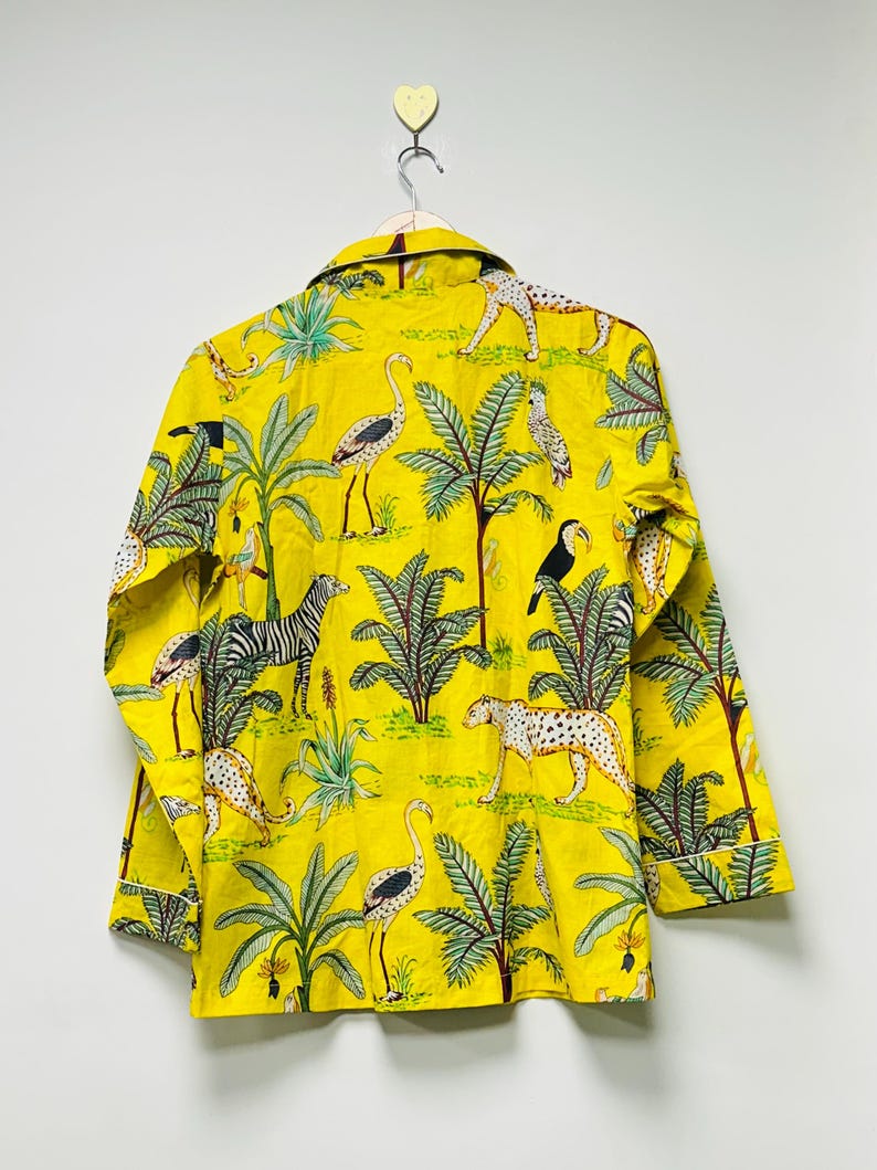 Puede incluir: Camisa amarilla con botones con estampado de tem&aacute;tica selv&aacute;tica. La camisa presenta cebras, leopardos, tucanes y flamencos entre palmeras y otro follaje. La camisa est&aacute; colgada en una pared blanca.