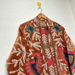 Puede incluir: Una chaqueta de terciopelo color &oacute;xido con bordados intrincados en crema, rosa intenso, azul oscuro y verde salvia. El dise&ntilde;o incluye motivos florales y de hojas en el frente y las mangas, con cuello alto y cierre de cremallera central.