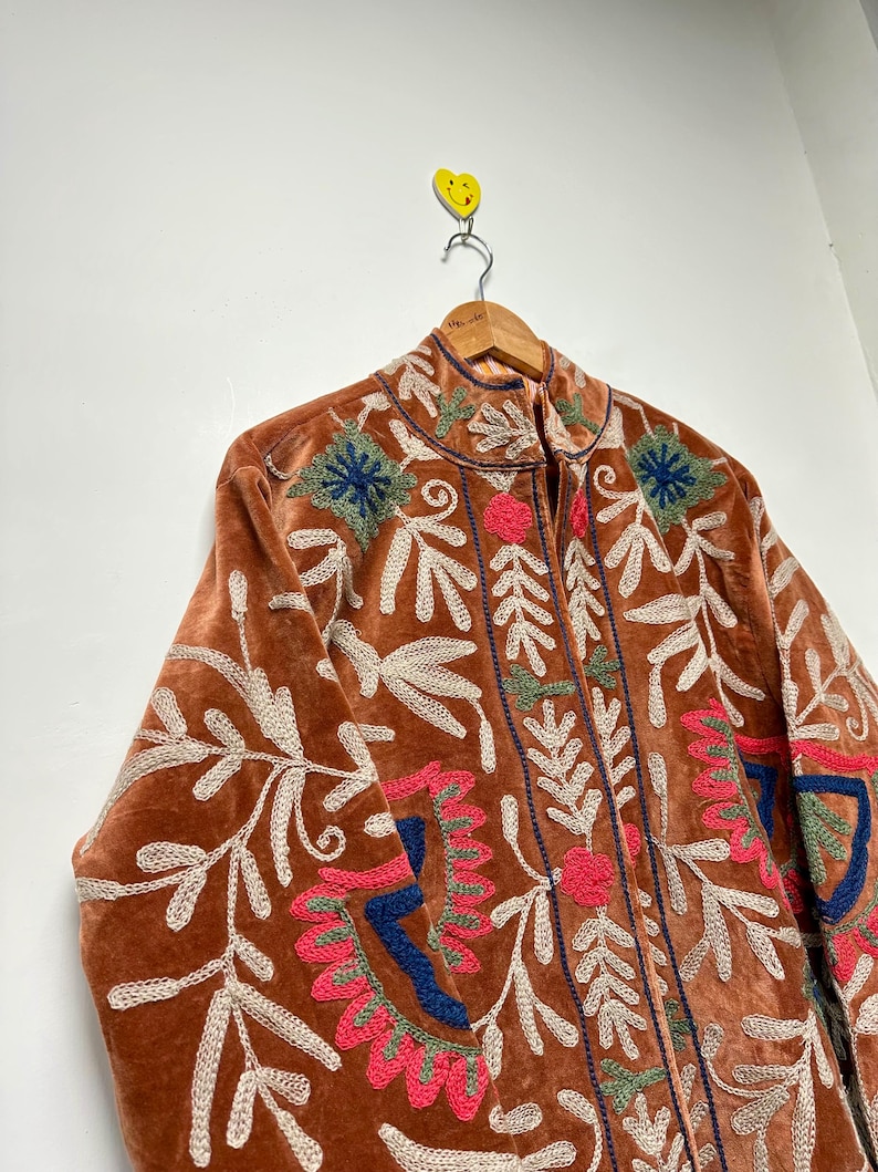 Puede incluir: Una chaqueta de terciopelo color &oacute;xido con bordados intrincados en crema, rosa intenso, azul oscuro y verde salvia. El dise&ntilde;o incluye motivos florales y de hojas en el frente y las mangas, con cuello alto y cierre de cremallera central.