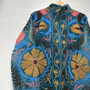 May include: A teal jacket featuring intricate embroidery in shades of mustard yellow, bright blue, dark brown, and soft pink. The design includes large floral motifs and scrolling vine patterns across the front and sleeves, with a high collar.