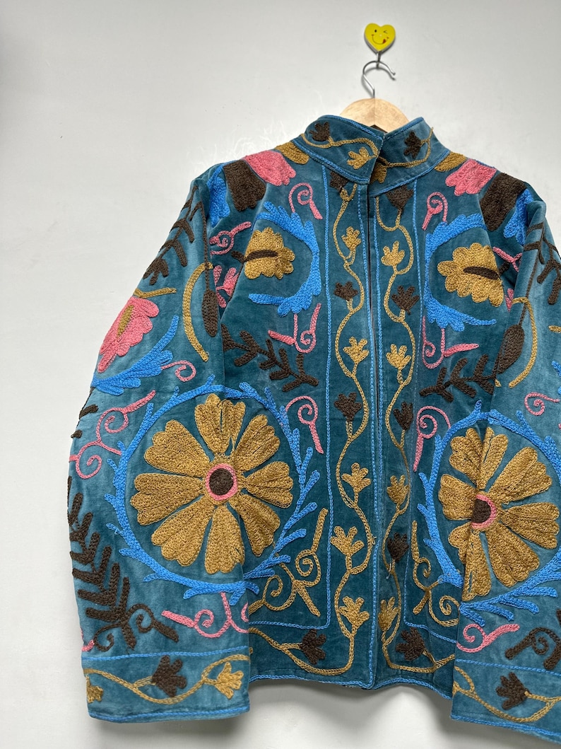 May include: A teal jacket featuring intricate embroidery in shades of mustard yellow, bright blue, dark brown, and soft pink. The design includes large floral motifs and scrolling vine patterns across the front and sleeves, with a high collar.