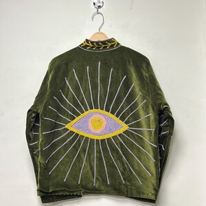 K&ouml;nnte beinhalten: Eine olivgr&uuml;ne Samtjacke mit einem gro&szlig;en gestickten Augenmotiv auf der R&uuml;ckseite. Das Auge ist in Lavendel-, Rosa- und Gelbt&ouml;nen mit strahlenden silbernen Strahlen dargestellt. Der Kragen zeigt ein gelbes, blattartiges Stickmuster.