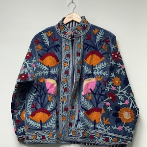 May include: A blue jacket featuring intricate floral embroidery in shades of orange, pink, red, and light blue. The garment has a high collar, long sleeves, and a front opening, with a visible striped lining visible along the inner edges.