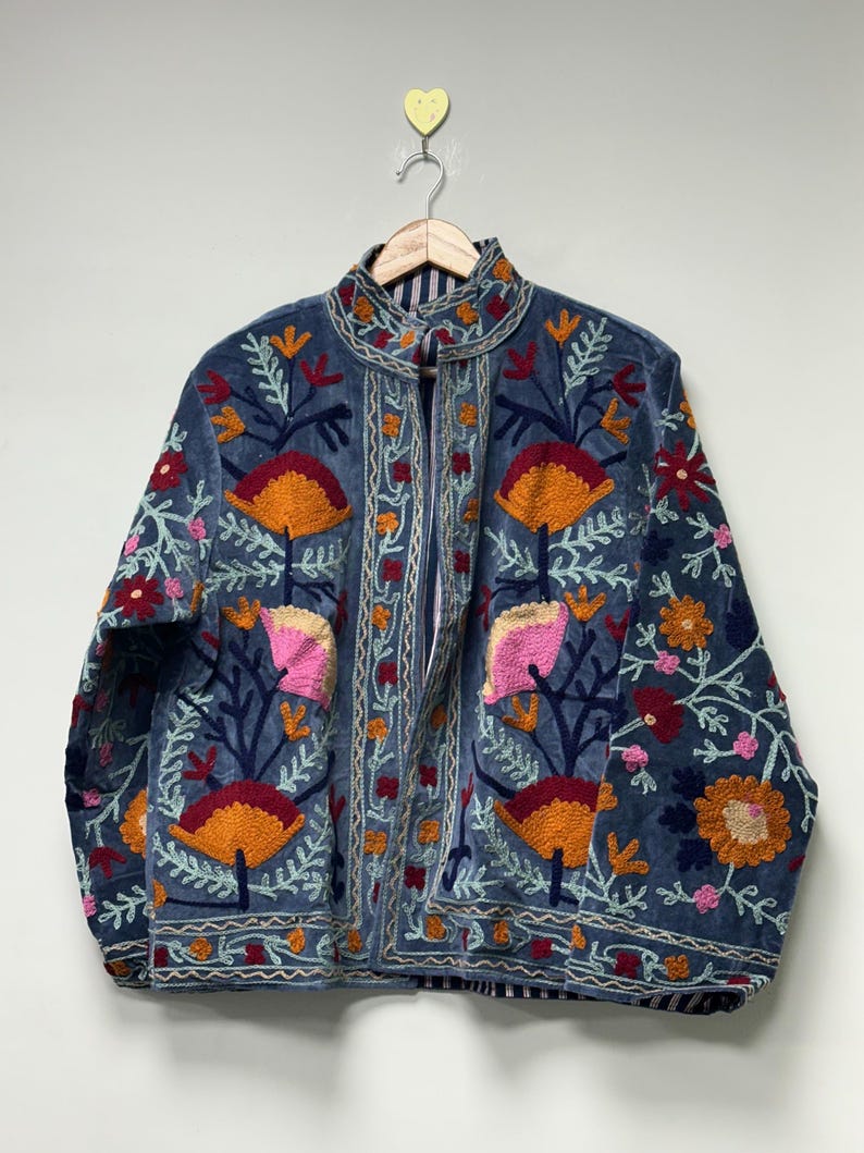 May include: A blue jacket featuring intricate floral embroidery in shades of orange, pink, red, and light blue. The garment has a high collar, long sleeves, and a front opening, with a visible striped lining visible along the inner edges.