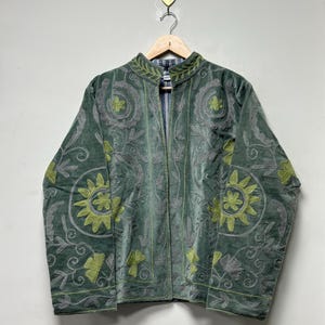 May include: A green velvet jacket with intricate floral embroidery in gray and lime green. The jacket has a mandarin collar and a striped lining. It is displayed on a wooden hanger.