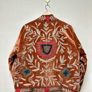 Puede incluir: Una chaqueta de terciopelo color &oacute;xido con intrincados bordados en crema, rojo, azul y verde. El dise&ntilde;o incluye patrones florales y de hojas sim&eacute;tricos, un motivo central en forma de escudo y detalles circulares decorativos en el dobladillo y el cuello.