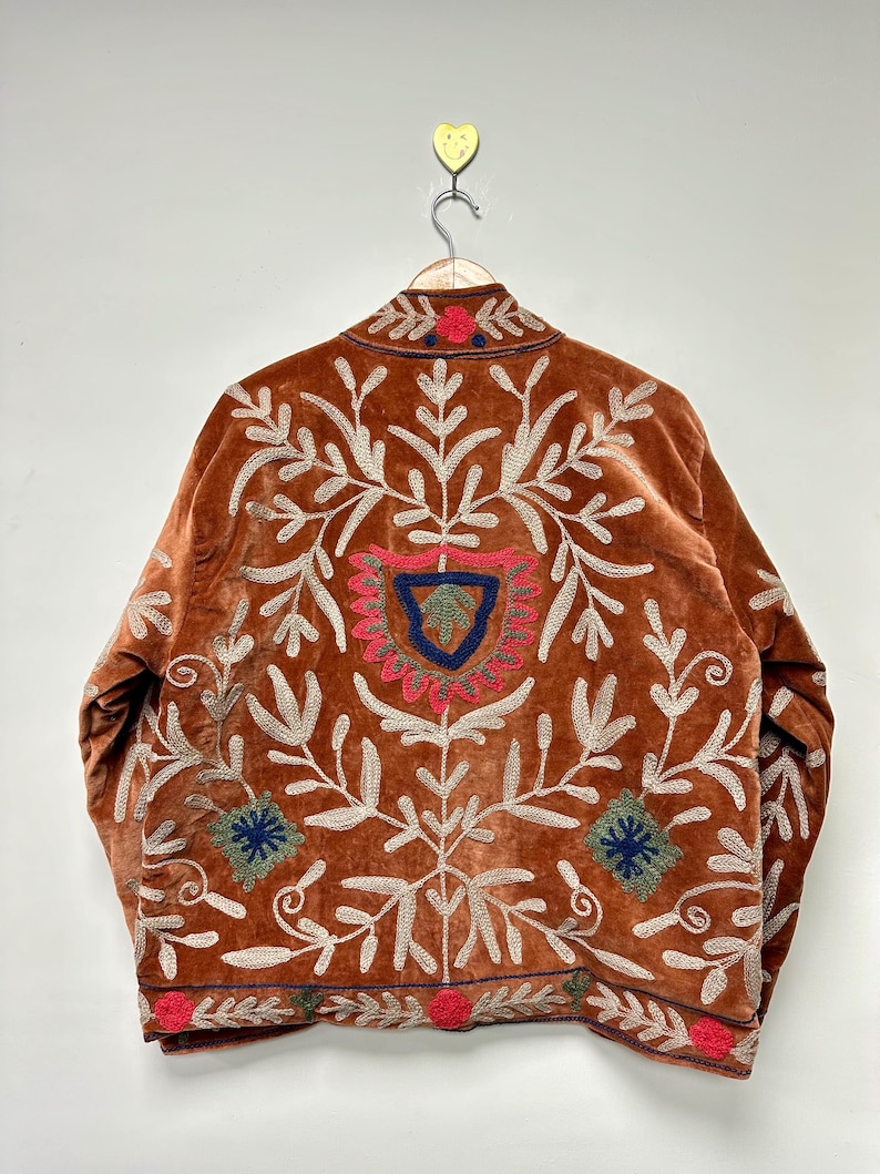 Puede incluir: Una chaqueta de terciopelo color &oacute;xido con intrincados bordados en crema, rojo, azul y verde. El dise&ntilde;o incluye patrones florales y de hojas sim&eacute;tricos, un motivo central en forma de escudo y detalles circulares decorativos en el dobladillo y el cuello.