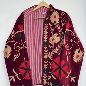 May include: A deep burgundy velvet jacket featuring intricate floral embroidery in shades of gold, red, and dark green. The garment has a stand-up collar and an open front, revealing a lining with a vertical pattern of thin red and white stripes.