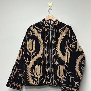 May include: A black jacket featuring long sleeves, a high collar, and an open front. The garment is decorated with intricate gold-coloured embroidery depicting stylised floral and leaf patterns across the front and sleeves on a dark fabric.