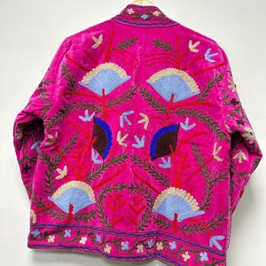 K&ouml;nnte beinhalten: Eine leuchtend fuchsiafarbene Jacke mit aufwendigen Stickereien aus hellblauen f&auml;cherf&ouml;rmigen Motiven, dunkelbraunen Akzenten und roten Zweigen. Das Kleidungsst&uuml;ck verf&uuml;gt &uuml;ber einen Stehkragen und eine dekorative Bord&uuml;re am Saum mit kleinen Blumenmustern.