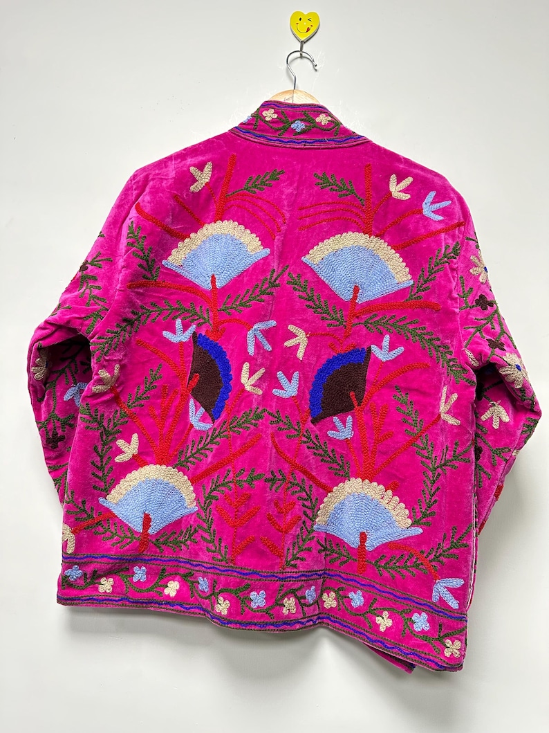 K&ouml;nnte beinhalten: Eine leuchtend fuchsiafarbene Jacke mit aufwendigen Stickereien aus hellblauen f&auml;cherf&ouml;rmigen Motiven, dunkelbraunen Akzenten und roten Zweigen. Das Kleidungsst&uuml;ck verf&uuml;gt &uuml;ber einen Stehkragen und eine dekorative Bord&uuml;re am Saum mit kleinen Blumenmustern.