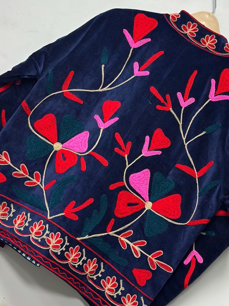 Op de afbeelding: Een marineblauw fluwelen jasje met levendig bloemenborduursel in tinten rood, roze en donkergroen. Het ontwerp bevat gestileerde bloemmotieven en kronkelende stengels, met een decoratieve geborduurde rand langs de onderzoom.