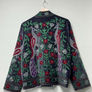 May include: A dark gray velvet jacket featuring intricate embroidery of two peacocks facing each other, surrounded by red flowers and green leafy vines. The design includes silver and purple accents on the peacock feathers and a patterned border.