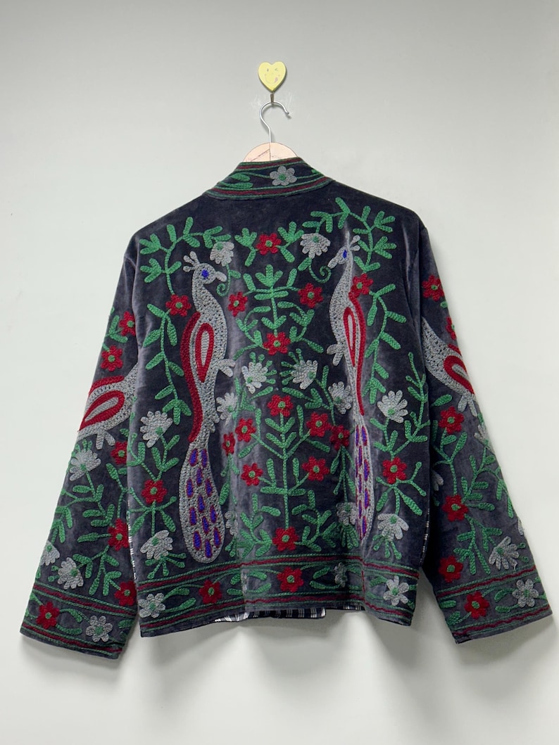 May include: A dark gray velvet jacket featuring intricate embroidery of two peacocks facing each other, surrounded by red flowers and green leafy vines. The design includes silver and purple accents on the peacock feathers and a patterned border.