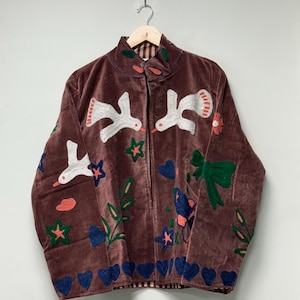 May include: A brown velvet jacket with embroidered white birds, flowers, stars, and hearts. The jacket has a high collar and a zip closure. The lining is a red and white plaid pattern.
