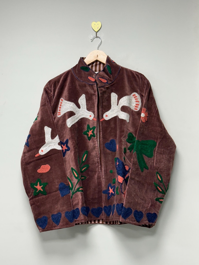 May include: A brown velvet jacket with embroidered white birds, flowers, stars, and hearts. The jacket has a high collar and a zipper closure. The lining is a red and white plaid pattern.