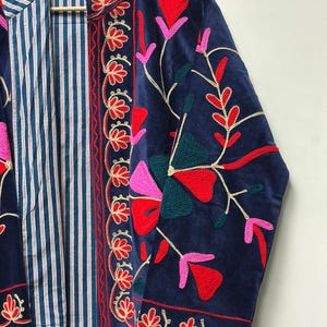Op de afbeelding: Een marineblauw fluwelen jasje met levendige bloemenborduursels in rood, roze en donkergroen. Het jasje heeft een opstaande kraag en een contrasterende binnenvoering met een blauw-wit verticaal streeppatroon op een lichte achtergrond.