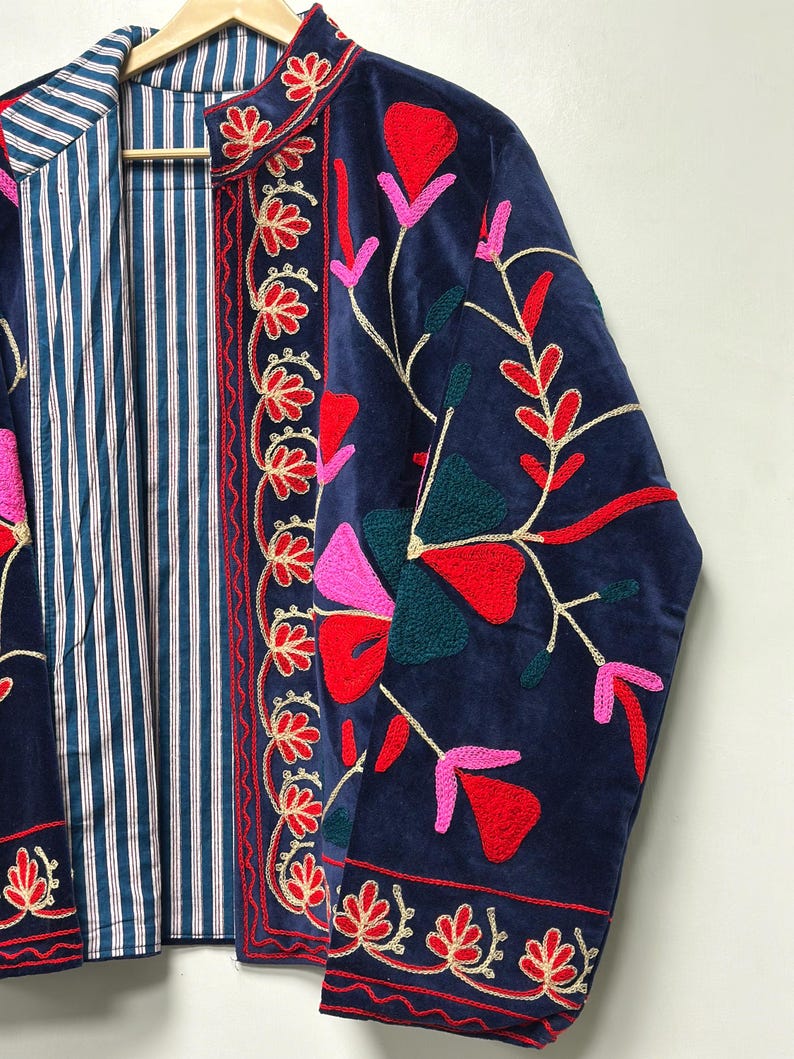 Op de afbeelding: Een marineblauw fluwelen jasje met levendige bloemenborduursels in rood, roze en donkergroen. Het jasje heeft een opstaande kraag en een contrasterende binnenvoering met een blauw-wit verticaal streeppatroon op een lichte achtergrond.