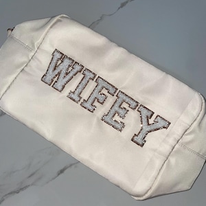 Può includere: Pochette cosmetica color crema con la scritta "WIFEY" ricamata in stile collegiale. Le lettere sono bordate in un marrone glitterato, con un riempimento bianco strutturato. La borsa è rettangolare con chiusura a zip.