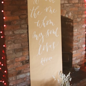 May include: A brown paper scroll with white calligraphy that reads "I have found the one whom my soul loves". The scroll is hanging on a brick wall with red Christmas lights.