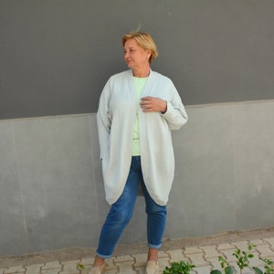 May include: A woman wearing a light gray cardigan over a light green shirt and blue jeans. She is standing in front of a gray wall.