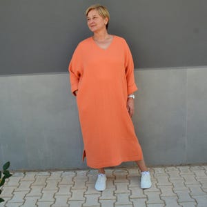 May include: A coral orange, long-sleeved, v-neck dress with a relaxed fit. The dress has a slightly gathered look and a side slit. The model is wearing white trainers.